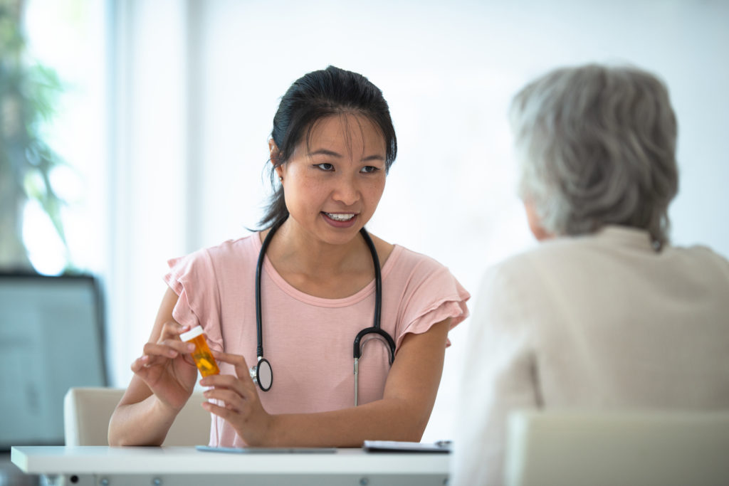 A female Asian doctor meets with her elderly patient to go over her new prescription medication. The doctor is going over the instructions for taking the medication and the possible side effects, as she holds the medication out in her hands. The woman is dressed casually and is looking at the doctor attentively.