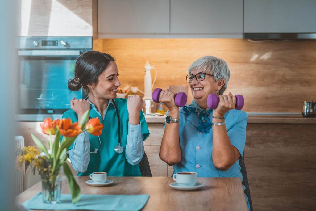 An elderly woman lifts light weights while sitting at a table with a medical professional sitting next to her